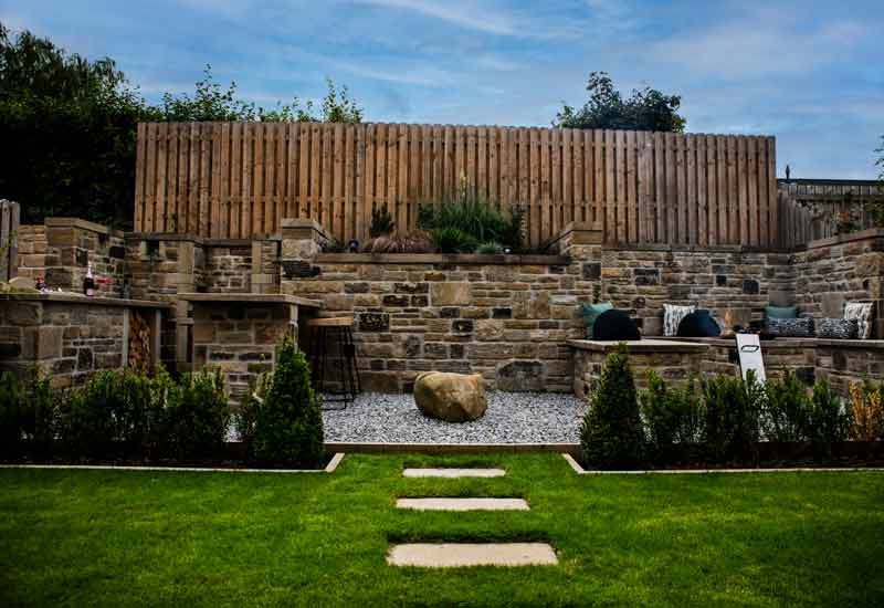 Grass area with stone stepping stones leads to an outdoor kitchen area slightly covered by bushes.