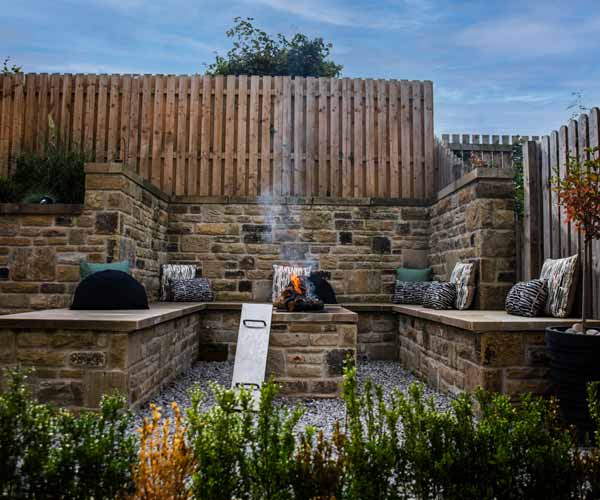 Squared off seating area surrounds a blazing fire pit with bushes in the foreground.